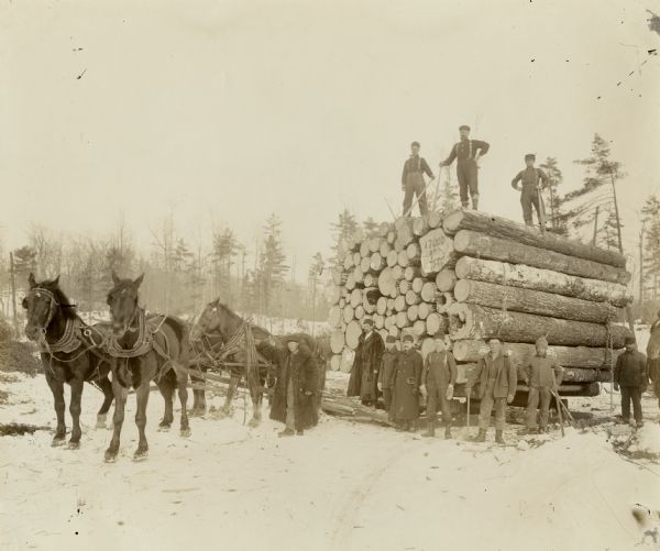 Eleven men are posing on or near a tall stack of logs on a sled. Some of the men are holding cant hooks. The logs are being hauled by a team of four horses. One log appears to have writing on its end, which says: "17000 Last Load 189(?)."