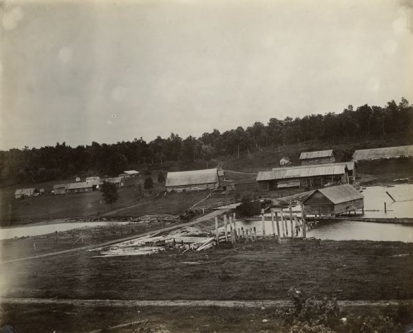 View of a lumber camp, with several buildings in the background. A bridge spans a river near the dam site.