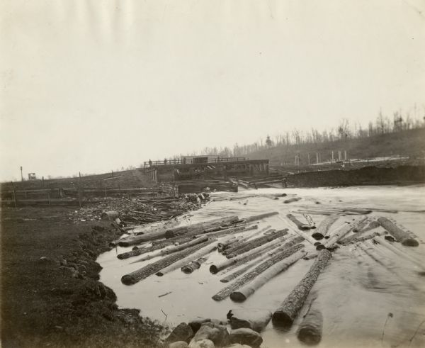 View of logs gathering in water just past a sluicing gate. Three people are standing on top of the dam.