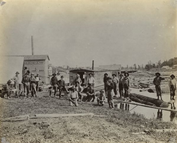 View of several men standing and kneeling in front of mill buildings. There are logs floating near the sluice gate.