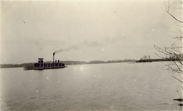 View across water towards a tow boat on the lake. Two men are standing on the top of the boat near a chimney spewing smoke.