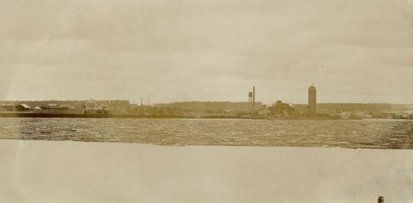 View across water towards logs floating along the lake. Several buildings, including a water tower and a silo, are on the opposite shoreline.