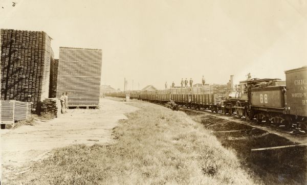 View of a trainyard, with several carloads of lumber on the train. Men are standing on the lumber. Additionally, several large stacks of lumber are in the yard, and two men are standing near them.