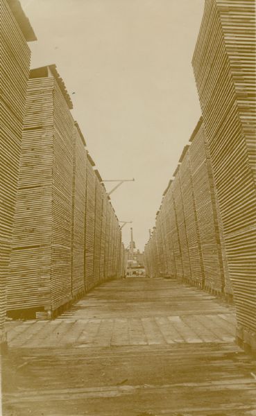 View along a boardwalk lined with tall stacks of lumber on both sides. Mill buildings are in the distance.