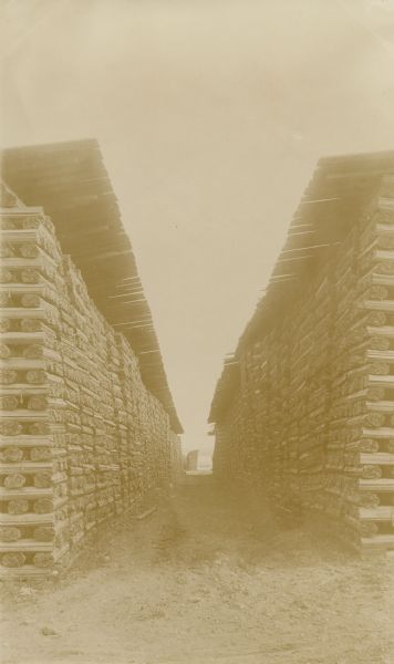 View between two rows of stacked lumber, which is slatted and bundled under a wood roof.