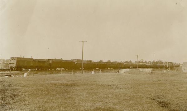 View across field towards a train loaded with slatted lumber piled into train cars. Telephone poles are along the railroad tracks.