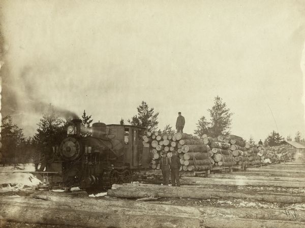 Several men are posing among logs on the ground near a train engine, which has continuous track (caterpillar) wheels and is pulling a load of logs on sledges. There is a man standing in the doorway of the locomotive, and a man, the steersman, is sitting on the front of the engine behind a steering wheel above the sled tracks. Another man is standing on the stack of logs chained together behind the engine. There is a total of about nine sledges behind the engine. In the right foreground is a log that has a mark that has been darkened on the photograph. Caption reads: "Early step in modernization. 'Cat' [A cat, or caterpillar, train] rolls horse & ox for hauling job. This bark-mark [arrow pointing to aforementioned mark] may be a clue to date and name of this later day [sic] operation."