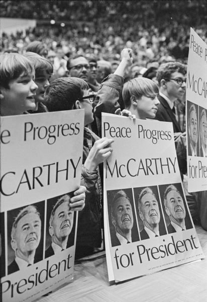 A group of teenage boys are in front of large crowd at a Democratic presidential candidate rally for Sen. Eugene McCarthy. They are holding large posters which read: "Peace Progress McCarthy for President." The rally was held at the Dane County Memorial Coliseum, where some 18,000 persons attended.