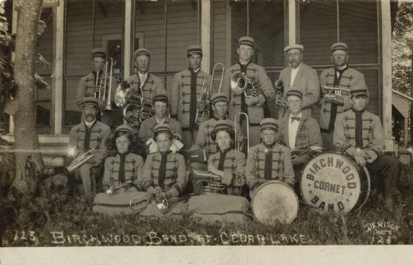 Outdoor group portrait of a band, wearing their uniforms and holding their instruments. The band is posed on the lawn in front of the porch of a house, and a drum on the right has a sign that reads: "Birchwood Cornet Band." Caption reads: "Birchwood Cornet Band at Cedar Lake."