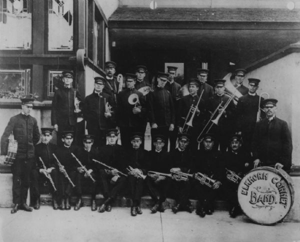 Outdoor group portrait of a band. The men are posing in their uniforms and are holding their instruments. A drum on the right has a sign that reads: "Elkhorn Cornet Band." A caption on the photograph mentions that the band is posing in front of the Lake Geneva Hotel, which was designed by Frank Lloyd Wright.
