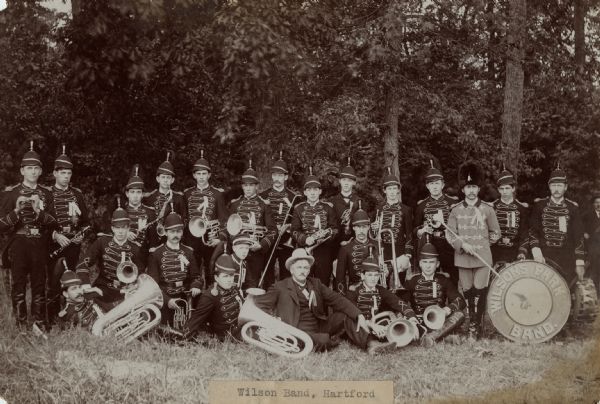 Group portrait of a band posing with the instruments, in uniform. The band is posing in front of a forest. The sign painted on the drum reads: "Wilson's Park Band." Caption reads: "Wilson Band, Hartford."