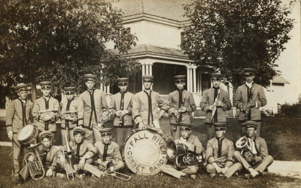 Outdoor group portrait of a band posing on the lawn. There is a buildings with a porch in the background. The men are wearing uniforms and holding their musical instruments. In the center a drum has a sign that reads: "Fall River Cornet Band."