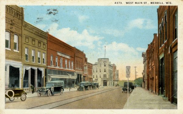 Colorized postcard view along Main Street, with several storefronts. Cars are parked along both sides of the street, and streetcar tracks are running down the center of the street. Caption reads: "West Main St., Merrill, Wis."