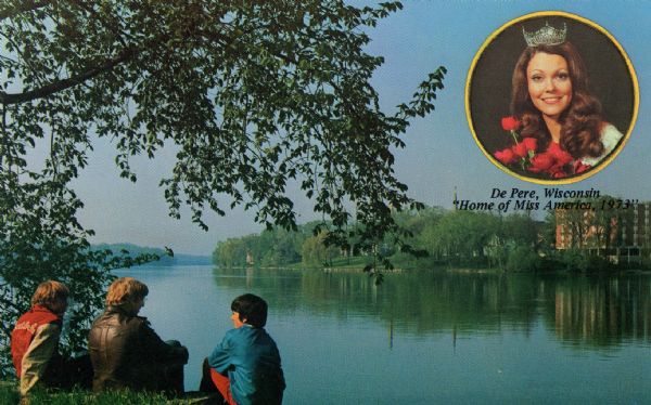 View towards the shoreline of a river, with three boys sitting on the bank in the foreground. There is a building among trees on the opposite shoreline. An inset image at top right is of a smiling woman holding roses and wearing a crown above the caption: "De Pere, Wisconsin, Home of Miss America, 1973." Caption on back of postcard reads: "This beautiful view is of the Fox River, which flows northward through De Pere, Wis., pop. 13,000. Designated by U.S. Congress as 'America's No. 1 Small City' because of it's [sic] enviable voting record, De Pere is also the home of Terry Anne Meeuwsen, chosen 'Miss America, 1973.'"