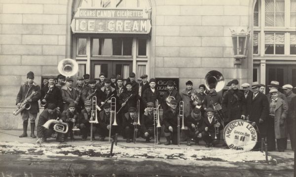 Members of a band posing outdoors for a group portrait with their musical instruments. The band members are wearing coats and hats, and a few are wearing men's flannel shirts. A drum is labeled "American Legion Band, Green Bay No. 11." They are standing outside a building which has a sign above the entrance advertising "Cigars, Candy, Cigarettes, Ice Cream."
