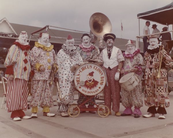 Group of seven men posing in clown costumes and holding musical instruments. A drum on a wheeled cart has a cartoon representation of a clown, and is labeled "Mel's Circus Clown Band, Sheboygan." Behind the group  on the right is a roofed platform with three men standing on it. 