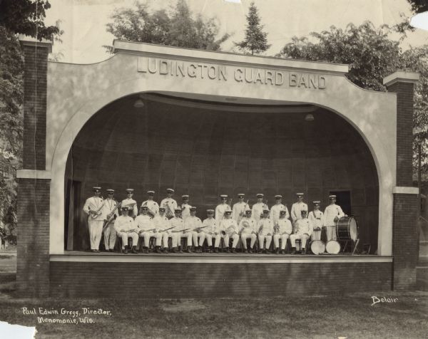 View across lawn towards band members posing in their uniforms and holding their musical instruments. The group is sitting and standing on stage under a band shell that bears the name "Ludington Guard Band." The caption reads: "Paul Edwin Gregg, Director, Menomonie, Wis."
