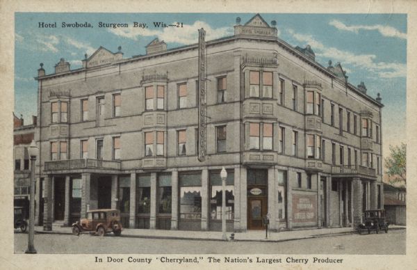 Color postcard showing a three-story building, which has several signs indicating it is the Hotel Swoboda. Three cars are parked outside the building, and neighboring buildings are partially visible. Streetlights are on the sidewalks. Caption reads: "In Door County 'Cherryland,' The Nation's Largest Cherry Producer."
