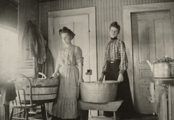 Two women are standing next to wash basins indoors. Behind them are two closed doors and a coat rack. In the foreground is a teakettle. Caption reads: "Ella Tillstrom (L) & Emma Bowman (R) doing the washing in Tillstrom Kitchen in Seymour Wisconsin."