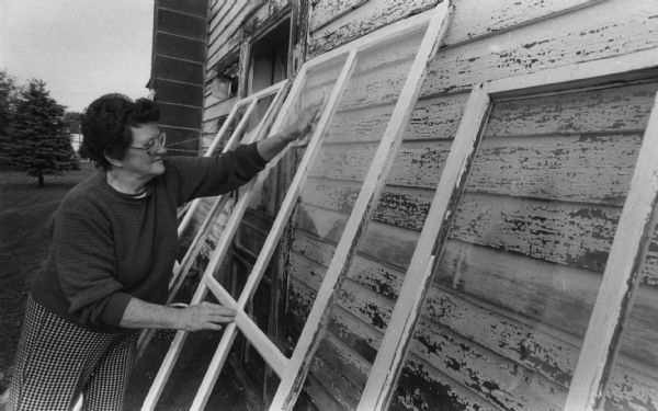 A woman is washing several windows that are leaning against the side of a building. Front of image is captioned: "Joyce Rhoades of Merrillan, WI. washed storm windows." Caption reads: "Getting ready for winter, Joyce Rhoades washed storm windows before putting them on her house in Merrillan."