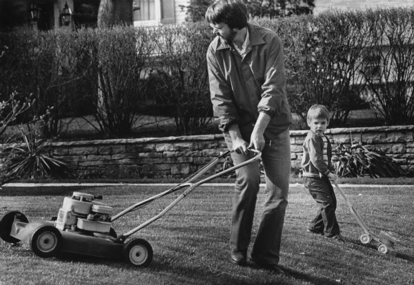 A man is mowing a lawn with a gas-powered mower. Behind him is a boy pushing a toy lawn mower. This photograph has two accompanying captions. The April 24, 1983, caption reads: "LITTLE MOWER, BIG HELPER — Vince, 3, maneuvered his toy mower while he watched his father, Mark Rutzinski, use a power mower on their Waukesha lawn."
The April 25, 1983, caption reads: "LIKE FATHER... — Vince, 3, tried to maneuver his toy lawn mower just like his father, Mark Rutzinski, operated the big power unit. They were working on the lawn of their home in Waukesha."