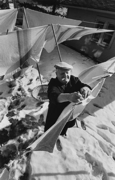 Overhead view of a man hanging clothes on a clothesline, while holding a clothespin in his mouth. Snow is on the ground. Caption reads: "LIFE IS A BREEZE — Nearly everyone has found a way to take advantage of the spring-like weather, including Frank Zaydell, 5709 W. Philip Pl., who hung his laundry out to dry Monday. More of the pleasant temperatures are expected Wednesday."