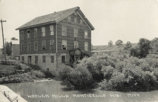 View across water towards the Historic Monticello Woolen Mill. There are many windows on the mill building, and some of them are open. Other buildings are behind the mill, and in the far background is a hill. Caption reads: "Woolen Mills, Monticello, Wis."