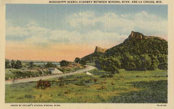 Colortone view towards a bend in a highway. The Mississippi River is in the far distance. To the right of the highway is a large field with a fence running through it, and partially wooded hills and bluffs are in the distance on the right. Caption reads: "Mississippi Scenic Highway Between Winona, Minn. and La Crosse, Wis."