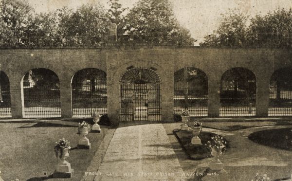 Slightly elevated view of the front gate at the Wisconsin State Prison, taken from inside the prison grounds. In the foreground is the walkway to the gate lined with plants in large planters, which are shaped like vases on pedestals. Caption reads: "Front Gate, Wis. State Prison, Waupun, Wis." The postcard was sent to Mr. Elmer Kruschke of Burnett, Wis, from M. Anithor [sp?], who wrote: "This is where they shut the bad boys up."