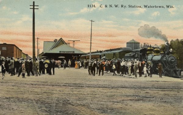 Tinted postcard view of crowds of people standing near a railroad train on the far right, and a train station in the center with a sign that reads: "Watertown." There are railroad cars on tracks on the left, telephone poles are along the platform, and a water tower is on the far right. Caption reads: "C. & N. W. Ry. Station, Watertown, Wis."