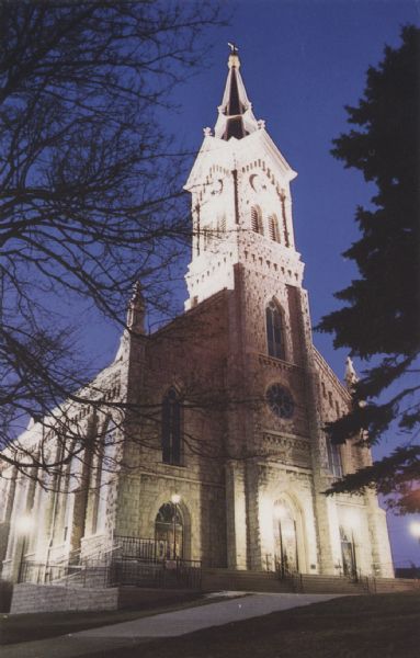 Night view of St. Mary's Church, showing the tower and front of the church. Streetlights and floodlights around the church illuminate the facade. The rear of the postcard gives the address: "St. Mary's Parish, 430 N. Johnson Port Washington, WI 53074 (414) 284-5771." 