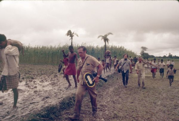 A film crew is carrying film equipment and walking in a muddy field, along with men, women and children. Palm trees are growing near a field of tall plants.