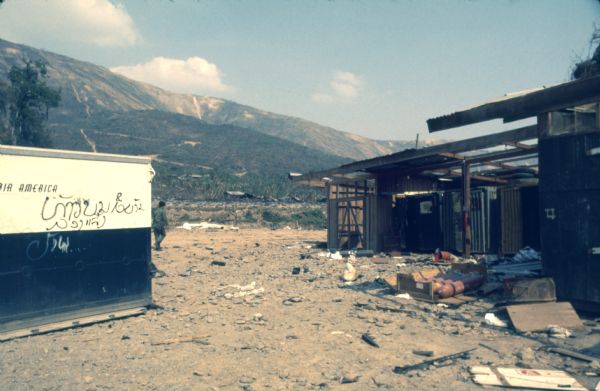 View of an Air America shipping container with graffiti on it. There are other buildings on the right with debris on the ground in front of them. A man is walking in the background, and in the far distance is a mountain.