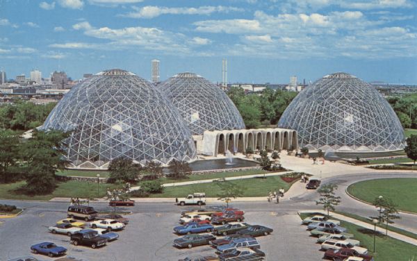 Elevated view of the three glass domes at the Mitchell Park Conservatory. Cars are parked in the parking lot and along the driveway, and groups of people are walking toward the conservatory. The Milwaukee skyline is in the distance. Caption on rear of postcard reads: "The 'Man made wonder,' the Mitchell Park Conservatory in the Milwaukee skyline. The three domed Conservatory offers a Tropical, an Arid, and a Floral display. Nearly 600,000 visitors annually view the exotic flowering plants. The Sunken Garden is an added feature during the summer months. Conservatory located at South 27th and West Pierce Street. Open 9 A.M. to 9 P.M. daily and weekends, except Mondays open 9 to 5."