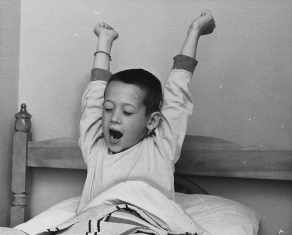 A boy is sitting up in bed and stretching with his mouth open, possibly in a yawn. Caption reads: "A good stretch after a long sleep and Robin Richards, 7, son of Mr. and Mrs. Stephen Richards, 9110 N. Range Line Rd., River Hills, was ready to play Monday. Robin returned home Sunday after having been the object of an extensive search Saturday in the Big Bay area of Michigan's Upper Peninsula. He wandered away from the home of his grandparents, where he was visiting. He was found eight hours later by a conservation officer two and one-half miles from the home of his grandparents. More than 100 volunteers, an airplane and bloodhounds took part in the search."