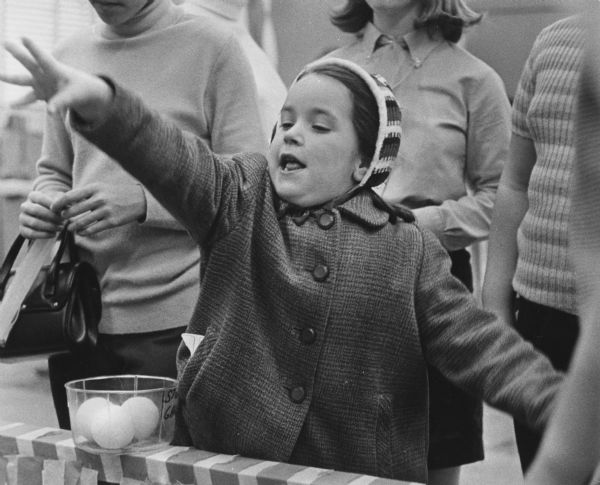 A girl wearing a coat and hat with her arm raised, as if she has just tossed something. In front of her is a clear bowl holding three small balls. Behind her are three people standing and watching. Caption reads: "ANOTHER WINNER - At the northwest branch of the YMCA Saturday, Lynne Marie Woods, 6, stepped up to the line and successfully tossed a little ball into a circle to win a prize. Lynne Marie, who lives at 4437 N. 66th St., took part in an afternoon Y carnival."