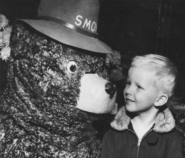 A boy is smiling and looking at the mascot, Smokey the Bear. Caption reads: "Nov. 1958. Smokey the Bear (James Coleman, U.S. Forester) and Thomas Smagalski." 