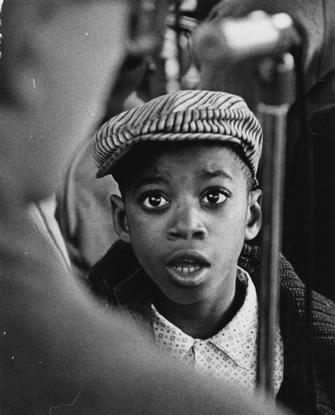 A boy is looking at a person in the foreground, who is out of focus. Another object — possibly a microphone stand — is also in the foreground and out of focus. Caption reads: "Robert Alexander, 9, of 1215 W. Hadley St., joined the singing as he watched Sams' guitar playing."