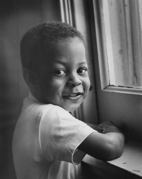 A boy is standing and resting his arms on a windowsill and smiling while looking over his shoulder towards the camera. Caption reads: "Kevin Stark, 2, of 2449 N. 11th St., spent the night at the county children's home because of a mix-up in family communications. A passer-by noticed him crying and called police Monday evening. Actually, he was outside the house next door to his home. Police canvassed the neighborhood asking if a child was missing. Kevin's grandmother, who had been caring for him, told police that he was not missing. She believed that he was still with his great-grandmother, living a block away, where he had been taken earlier by his sister. Both of Kevin's parents were working at the time."