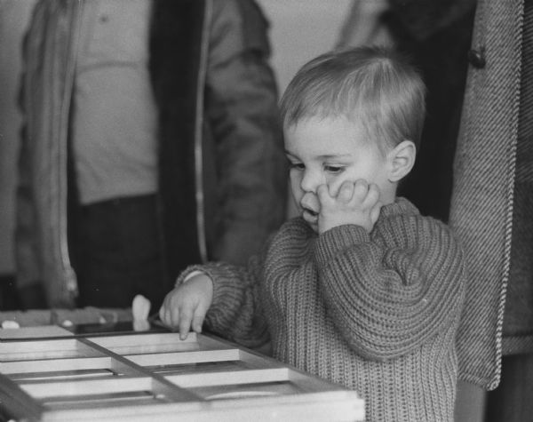 A young boy is scrunching up his cheek with one hand while pointing to something in a frame on a table. Adults are standing in the background (faces out of frame). Caption reads: "His courage gathered, Mark concentrated on forms on a table. He will be eligible for entrance to the academy [Elmbrook Montessori Academy, Milwaukee] when he is 2 1/2 years old."