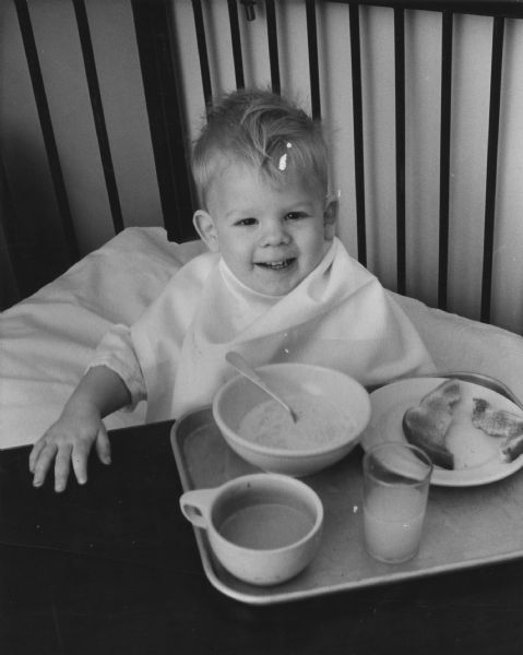 A boy is smiling and sitting up in a bed. He has one hand on a bedside table. On the table is a tray with a bowl of cereal, a mug, a glass, and a plate with toast. The boy is wearing a napkin as a bib. Caption reads: "A wandering boy, Jerome Mrozak, 23 months, 1335 S. 21st St., smiled over his breakfast at Johnston municipal hospital Saturday. Jerome's parents, Mr. and Mrs. Kenneth Mrozak, say he had just learned to open doors. He opened one at about 6:30 a.m. Saturday and strolled into an alley, wearing diapers and an undershirt. A woman saw him and took him to a near-by home. Police who were called took him to the hospital and checked the neighborhood to find where he lived. Jerome seemed to enjoy himself in the hospital, scampering in and out of bed over its high sides."