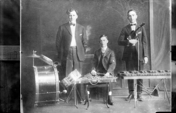 Three men are posing with musical instruments in front of a painted backdrop. One man is standing by a drum set on the left, a man is sitting in the center behind a musical instrument, and a man is standing on the right holding a violin, behind another instrument set up on a stand in front of him.