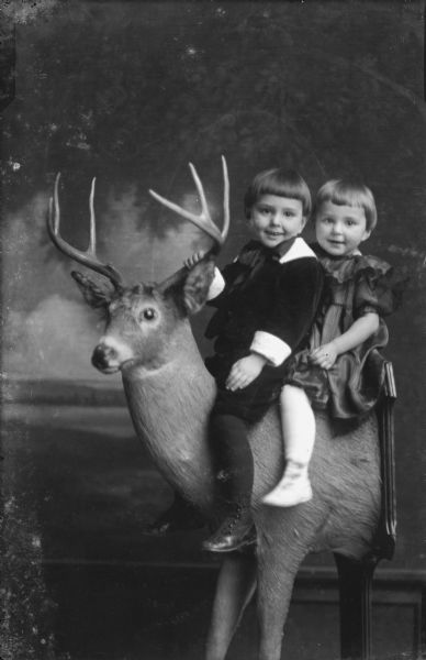 Studio portrait in front of a painted backdrop of two children, a boy and girl, sitting on the neck of a taxidermied deer with antlers. The front half of the deer is mounted on a stand and a plaque has been attached to the middle section as a backing.