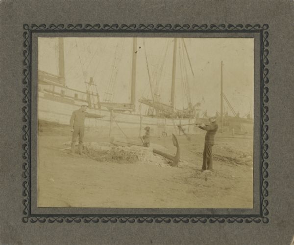 Two men are standing, one on the left with a pistol, and one on the right with a rifle, both aiming their weapons at a dog, which is sitting on its hind legs in the center on a brick wall. In the background is a ship named the <i>Hattie Hutt</i>, which was built in 1873 and caulked in 1888 for Louis Hutt, who might be one of the two men. The ship's port of hail was Chicago.