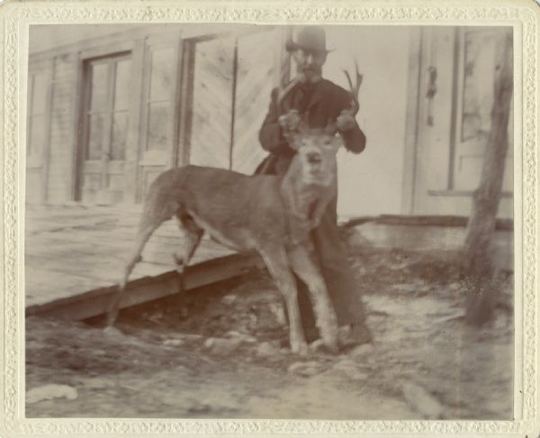 A man wearing a hat and suit is posing in front of a building and holding a buck, which appears to be dead. The man is holding the buck by its antlers and is turning its head to face the camera. Behind him is a building.