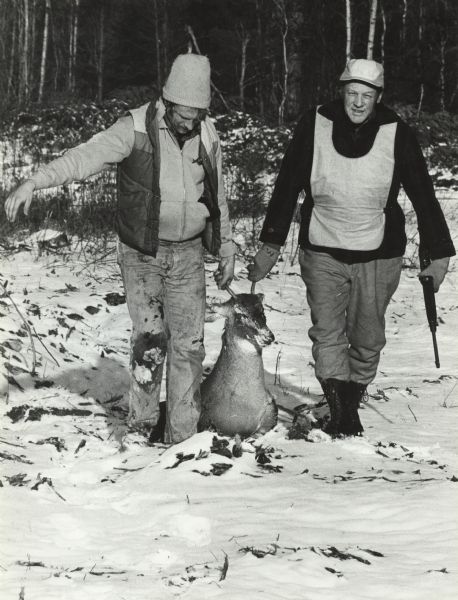 Two men are dragging a killed deer by its antlers out of a forest. One man is holding a rifle. Caption reads: "Dale Schneider, left, of Maribel, and Warren Wagner of Lark pulled a four-point buck out of the woods near Goodman Park. Schneider got the deer with a 20-gauge shotgun."