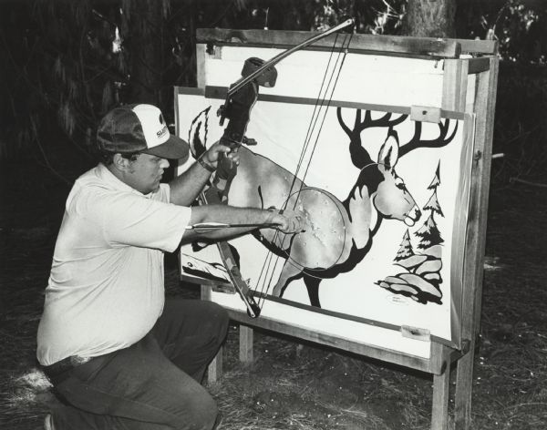 A man is kneeling and pulling an arrow out of a target, which has an illustration of a buck. The man is also holding a compound bow. Caption reads: "Russ Shepherd, assistant manager of the McMiller Shooting Center, retrieved a well-placed arrow from a target on the new bow-hunter field range at the facility near Eagle. Deer hunting season for Wisconsin archers begins Sept. 19."