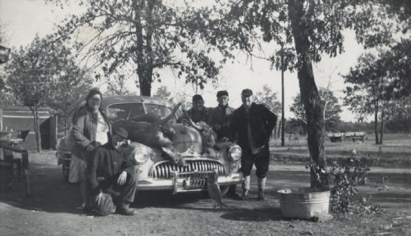 Four men and a woman are posing around a car, on which three killed deer are set on the hood of the car. A cat has its paws on the front of the car and is looking up at the deer. Caption reads: "Deer hunting about 1948 near New Miner. Taken by John E. Limbach, George Sterbens Farm."