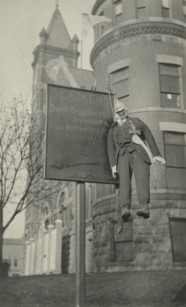 A sign outside a building reads "Slackers" and includes a list of names: David Englehardt of Browntown, and John Hoffmeister of Jords. The sign is credited to the Green County Council of Defense. Hanging from the sign is an effigy of a German dressed in a suit, with a fake Iron Cross and sheathed sword on its outfit.