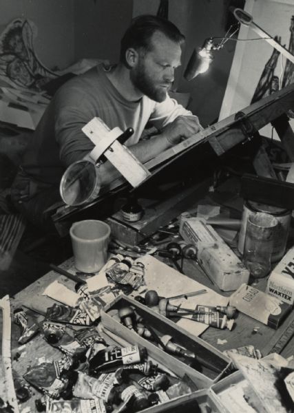 A man is working at a desktop easel, with an array of oil ink tubes and awls on the table next to him. A container of Curad bandages and a box of Northern Waxtex [sandwich bags] are on the right.
